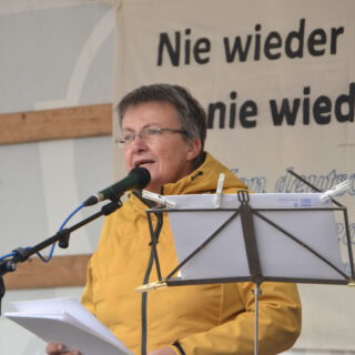 Eine Frau mit kurzen schwarzgrauen Haaren spricht in ein Mikro. Dahinter ein Transparent mit "Nie wieder Faschismus - nie wieder Krieg!" teilweise zu sehen.