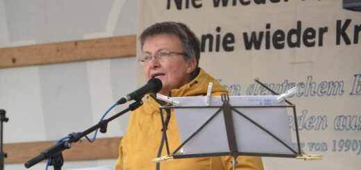 Eine Frau mit kurzen schwarzgrauen Haaren spricht in ein Mikro. Dahinter ein Transparent mit "Nie wieder Faschismus - nie wieder Krieg!" teilweise zu sehen.