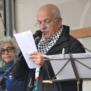 Ein älterer Mann mit grauem Haarkranz und Brille spricht in ein Mikro. Eine Frau mit grauen Locken schaut ihm ernst von hinten zu.