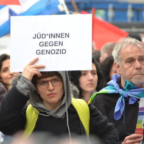 Eine junge Frau hält ein Demo-Schild hoch. Im Hintergrund dicht gedrängt Leute mit Fahnen.