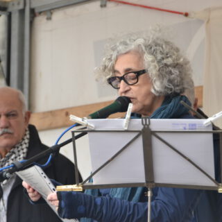 Eine Frau mit grauen Locken spricht in ein Mikro. Ein älterer Mann mit grauem Haarkranz und Brille schaut ihm ernst von hinten zu.