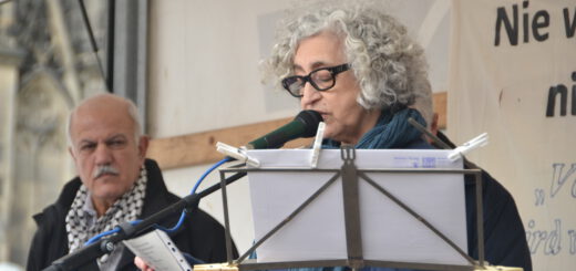 Eine Frau mit grauen Locken spricht in ein Mikro. Ein älterer Mann mit grauem Haarkranz und Brille schaut ihm ernst von hinten zu.