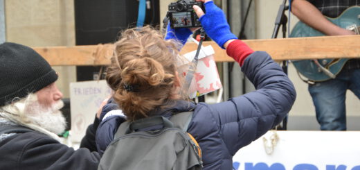Ein älterer Herr mit schwarzer Wollmütze und eine Frau mit hochgesteckten hellbraunen Haaren fotografieren Musiker auf einer Bühne.