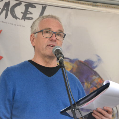 Ein Mann mit kurzen grauen Haaren und Brille in einem blauen Pulli spricht in ein Mikro. Im Hintergrund Teile eines Plakates mit "Peace!" und "La Paix!".