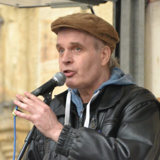 Ein Mann mit brauner Schlägermütze und schwarzer Lederjacke spricht in ein Mikro