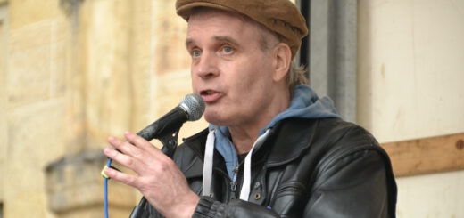 Ein Mann mit brauner Schlägermütze und schwarzer Lederjacke spricht in ein Mikro