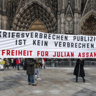 Vier winterlich gekleidete Personen tragen ein großes weißes Transparent mit schwarzer und roter Aufschrift. Im Hintergrund ein großes gotisches Kirchenportal.