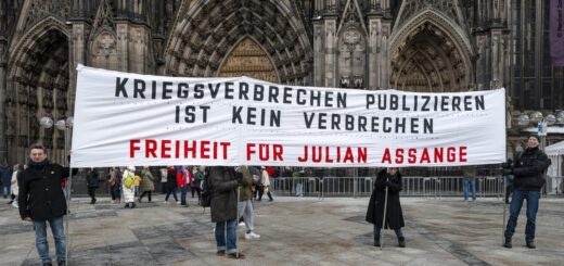 Vier winterlich gekleidete Personen tragen ein großes weißes Transparent mit schwarzer und roter Aufschrift. Im Hintergrund ein großes gotisches Kirchenportal.