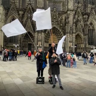 Zwei Frauen und ein Mann, darunter zwei auf leeren Getränkekisten stehend, schwenken große weiße Fahnen vor dem großen Portal eines gotischen Domes. Im Hintergrund Menschengruppen.