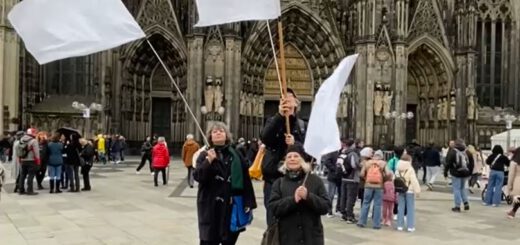 Zwei Frauen und ein Mann, darunter zwei auf leeren Getränkekisten stehend, schwenken große weiße Fahnen vor dem großen Portal eines gotischen Domes. Im Hintergrund Menschengruppen.
