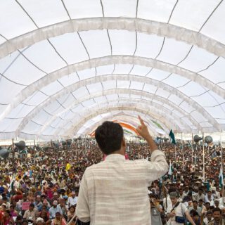 Ein Mann mit schwarzen kurzen Haaren spricht mit erhobenem Zeigefinger zu einer riesigen Menschenmenge, die in einem hellen weißen, tunnelförmigen Zelt steht.