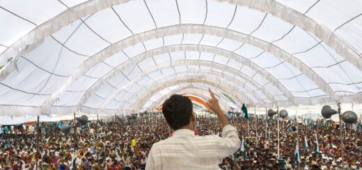 Ein Mann mit schwarzen kurzen Haaren spricht mit erhobenem Zeigefinger zu einer riesigen Menschenmenge, die in einem hellen weißen, tunnelförmigen Zelt steht.