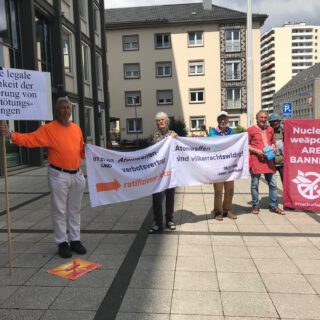 Vier Männer und eine Frau halten Transparente mit Aufschriften wie "Für eine legale Möglichkeit der Verhinderung von Massentötungsübungen" oder "Atomwaffenverbotsvertrag ratifizieren jetzt!" und "Atomwaffen sind völkerrechtswidrig". Im Hintergrund städtische Wohn- und Verwaltungsgebäude.