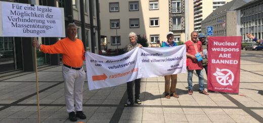Vier Männer und eine Frau halten Transparente mit Aufschriften wie "Für eine legale Möglichkeit der Verhinderung von Massentötungsübungen" oder "Atomwaffenverbotsvertrag ratifizieren jetzt!" und "Atomwaffen sind völkerrechtswidrig". Im Hintergrund städtische Wohn- und Verwaltungsgebäude.
