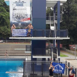 In einem Freibad hängt ein Großplakat mit einem Kriegsschiff und der Aufschrift "Karrieresprung" auf der Sprungplattform davor halten zwei Männer ein Banner mit der Aufschrift "Kein Werben fürs Sterben!". Darunter halten zwei weitere ein blaues Banner, auf dem das Logo der DFG-VK zu sehen ist: ein zerbrochenes Gewehr.