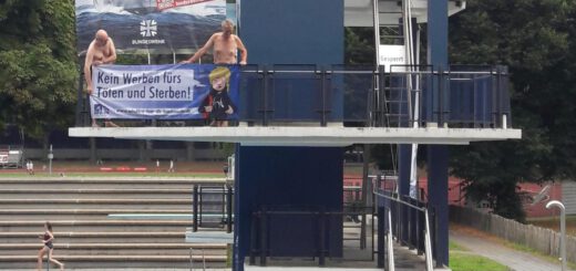 In einem Freibad hängt ein Großplakat mit einem Kriegsschiff und der Aufschrift "Karrieresprung" auf der Sprungplattform davor halten zwei Männer ein Banner mit der Aufschrift "Kein Werben fürs Sterben!". Darunter halten zwei weitere ein blaues Banner, auf dem das Logo der DFG-VK zu sehen ist: ein zerbrochenes Gewehr.