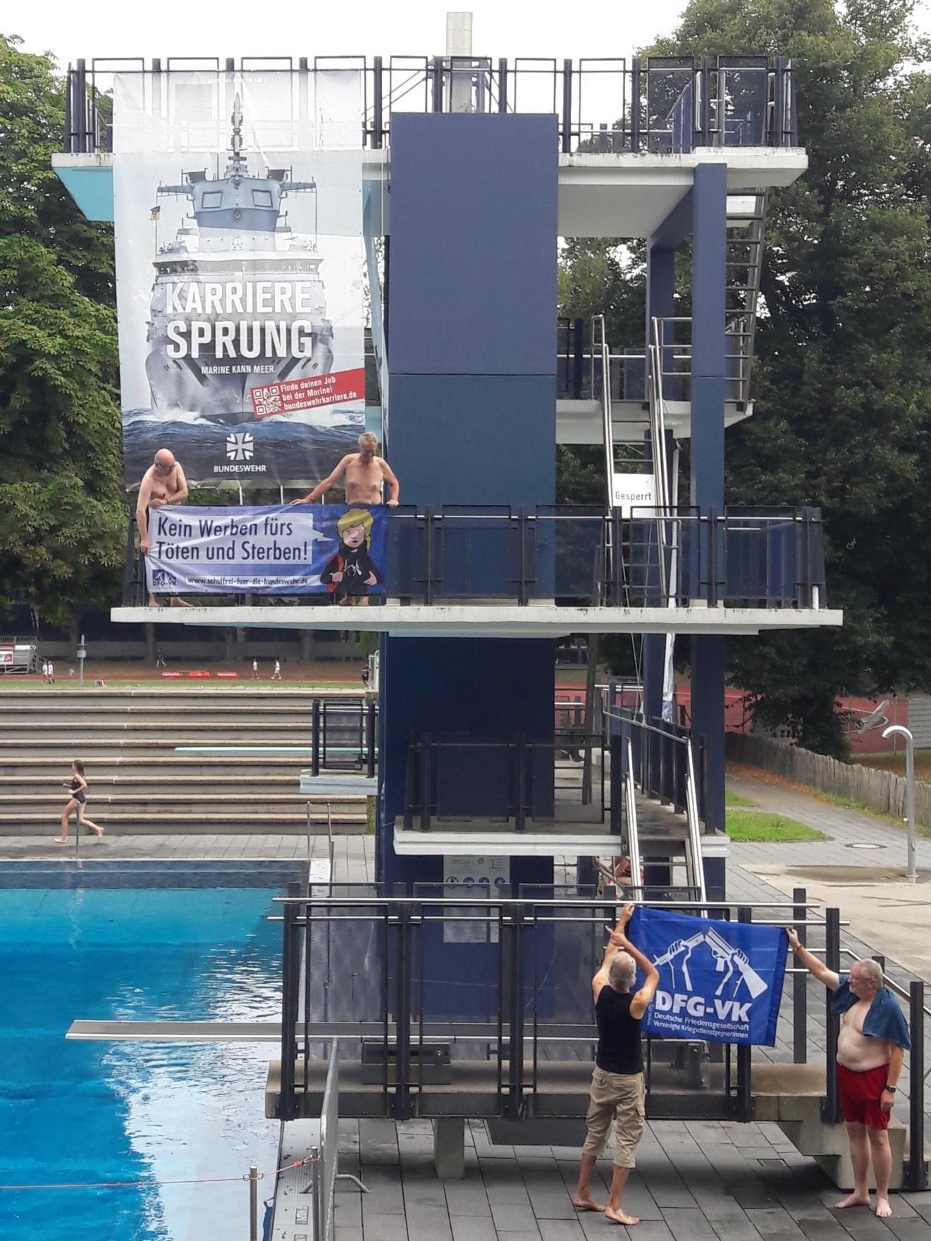 In einem Freibad hängt ein Großplakat mit einem Kriegsschiff und der Aufschrift "Karrieresprung" auf der Sprungplattform davor halten zwei Männer ein Banner mit der Aufschrift "Kein Werben fürs Sterben!". Darunter halten zwei weitere ein blaues Banner, auf dem das Logo der DFG-VK zu sehen ist: ein zerbrochenes Gewehr.
