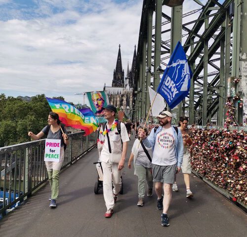 Eine Gruppe Menschen mit Fahnen läuft dem Betrachter entgegen. Im Hintergrund die zwei spitzen Türme des Kölner Doms.