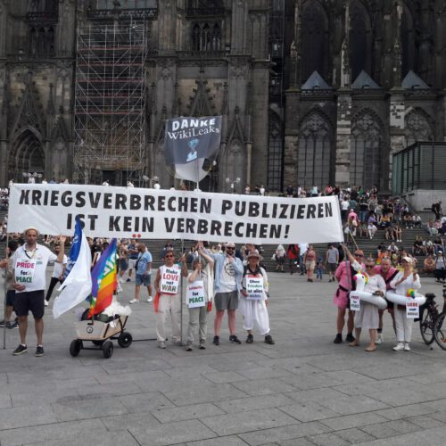 Eine Gruppe Menschen steht mit einem weißen Transparent mit schwarzer Schrift vor dem Hauptportal des Kölner Doms.
