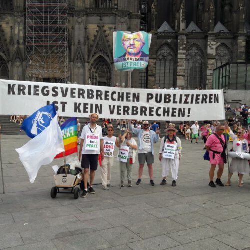 Eine Gruppe Menschen steht mit einem weißen Transparent mit schwarzer Schrift vor dem Hauptportal des Kölner Doms.